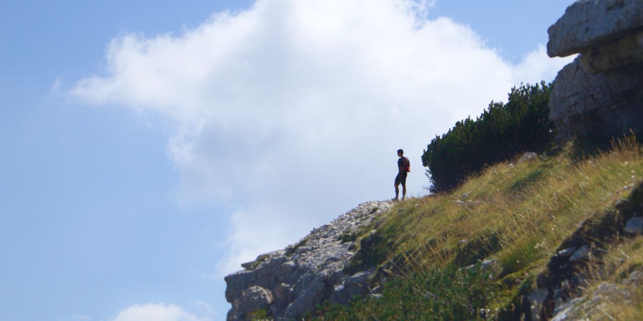 A lone hiker on a mountain ridge, seen from below against a large white cloud